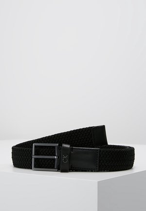 Ceinture en tissu noir tissé avec une zone de boucle en cuir noir portant le logo CK, affichée enroulée sur une surface blanche sur fond gris.