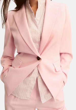 Donna che indossa un blazer su misura rosa chiaro e pantaloni coordinati su una camicia bianca con sottili righe verticali. Mani nelle tasche.