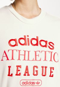 Roomkleurige sweatshirt met rood geborduurde tekst "adidas ATHLETIC LEAGUE" met logo, ronde hals en zachte textuur.