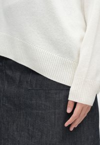 Λευκό πλεκτό πουλόβερ με ραμμένο μανίκι, επάνω από μια σκούρα γκρίζα τζιν φούστα με λεπτομέρεια πίσω τσέπης. Ορατό χέρι που στηρίζεται στη φούστα.