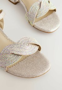 Sandalias plateadas brillantes con un upper texturizado y adornos de cuentas en un patrón ondulado. Diseño de puntera cuadrada y tacón bajo en bloque.