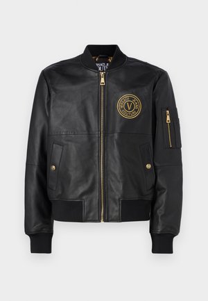 Giubbotto bomber in pelle nera con un logo circolare dorato sul petto, chiusura con zip frontale, polsini a coste e tasche laterali. Inclusi dettagli con zip dorata.