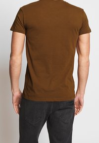Camiseta de algodón marrón con mangas cortas, cuello redondo y dobladillo recto. Llevada con jeans oscuros que tienen un parche de marca en la parte posterior.