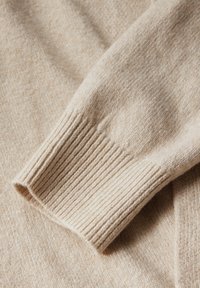 Fragment béžového pleteného svetr se žebrovaným manžetem, měkkou texturou a jemnými skvrnkami pro zvýraznění vizuálního dojmu.
