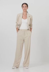 Frau steht, trägt einen beigen Anzug mit hochgekrempelten Ärmeln, weißen ärmellosen Top, beige weit geschnittene Hosen und Sneakers, Hände in den Taschen.