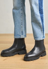Sorte lærres støvler med tykk såle og elastiske sidepaneler, kombinert med lyseblå jeans med en mørk stripe.