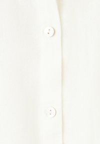 Gros plan d’un tissu blanc avec deux petits boutons ronds blancs cousus sur une patte verticale.