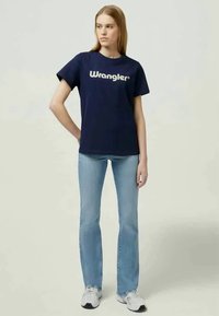 Tumedat sinise värviga lühikeste varrukatega T-särk, millel on valge "Wrangler" logo, koos helesiniste laienevate teksade ja valgete spordijalatsitega.