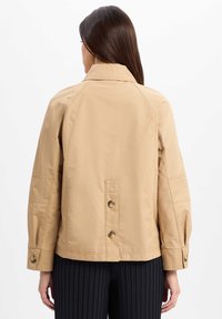 Khakifarbene Jacke mit Kragen, Raglanärmeln und Knopfdetails. Sie hat einen geraden Schnitt und eine glatte Textur, kombiniert mit plissierten schwarzen Hosen.