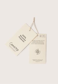 Étiquette en papier recyclé écru, composée de deux sections : l'une indiquant "100 % polyester recyclé", l'autre détaillant les pratiques durables avec une icône de globe.