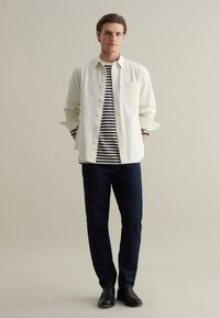Lichtbeige knoopshirt over een zwart-wit gestreept t-shirt, gecombineerd met donkere spijkerbroek en zwarte schoenen. Vlakke stoftexturen overal.