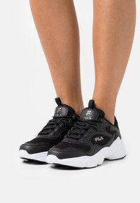 Chaussures de sport noires avec une tige en mesh et cuir synthétique, semelle intermédiaire blanche et lacets noirs. Présente le logo Fila sur la languette et sur le côté.