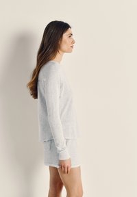 Pikkade pruuni juustega naine, kes kannab heledat halli pikkade varrukatega särk ja sobivaid lühikesi pükse, seisab profilvaates ühevärvilise beeži tausta ees.