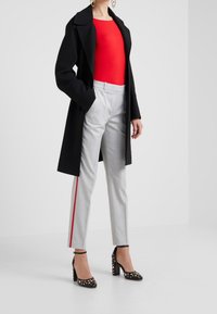Abrigo negro, blusa roja ajustada, pantalones grises a medida con raya lateral roja y tacones negros con tachuelas doradas. Materiales suaves y diseño estructurado.