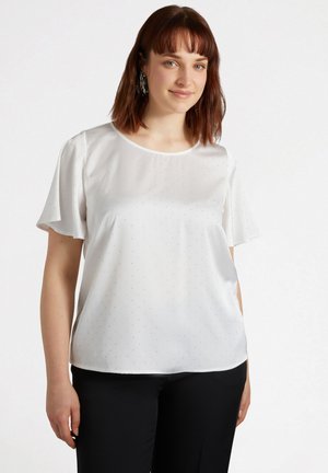Blouse blanche à manches courtes en tissu léger, avec un col rond et un motif à pois subtils. Un pantalon noir est porté en dessous.