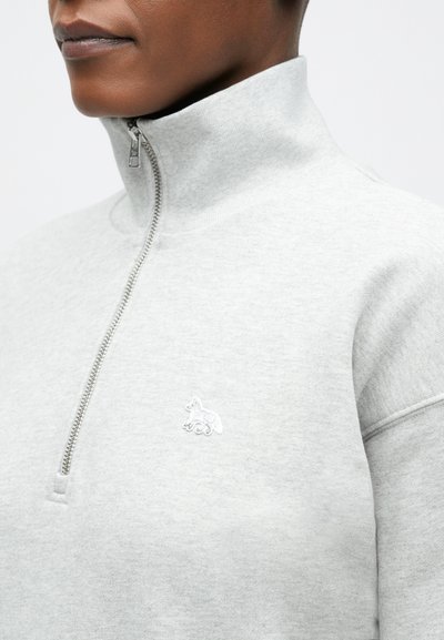 Svetlošedý mikina s vysokým golierom, polovičným zipsom a vyšitým logom líšky na hrudi. Mäkká, štruktúrovaná textília.