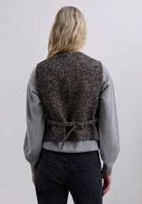 Chaleco marrón con patrón de espiga, de diseño ajustado. Presenta una cintura ceñida con un detalle de cinta, llevado sobre un suéter gris y jeans negros.