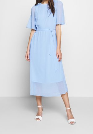 Femme portant une robe midi bleu clair avec des manches volantées et une ceinture nouée à la taille, associée à des talons blancs ouverts à l'avant avec bride à la cheville.