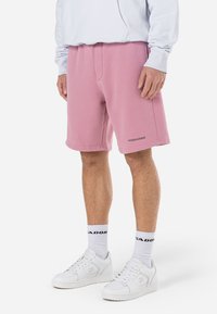 Lyserøde svedshorts lavet af blødt stof, med sidelommer og et logo ved kanten. Sammen med hvide sneakers og ankelsokker.