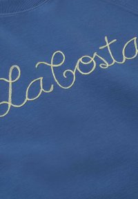 Tissu bleu avec une broderie jaune cursive épelant "Lacoste" sur toute la surface près du col côtelé.