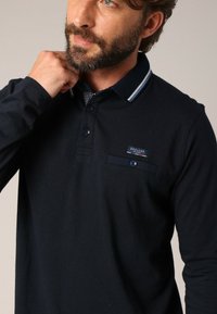 Polo à manches longues bleu marine avec un col contrasté, une poche poitrine et un patch logo. Le tissu a une finition texturée.