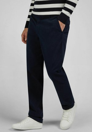 Homme portant un pantalon bleu marine, des baskets blanches et un t-shirt à manches longues rayé noir et blanc, une main dans la poche.