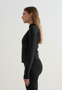 Černá sportovní bunda ve fitted designu, s vysokým límcem, dlouhými rukávy a kontrastním štepováním. Hladká textura, lehký materiál.