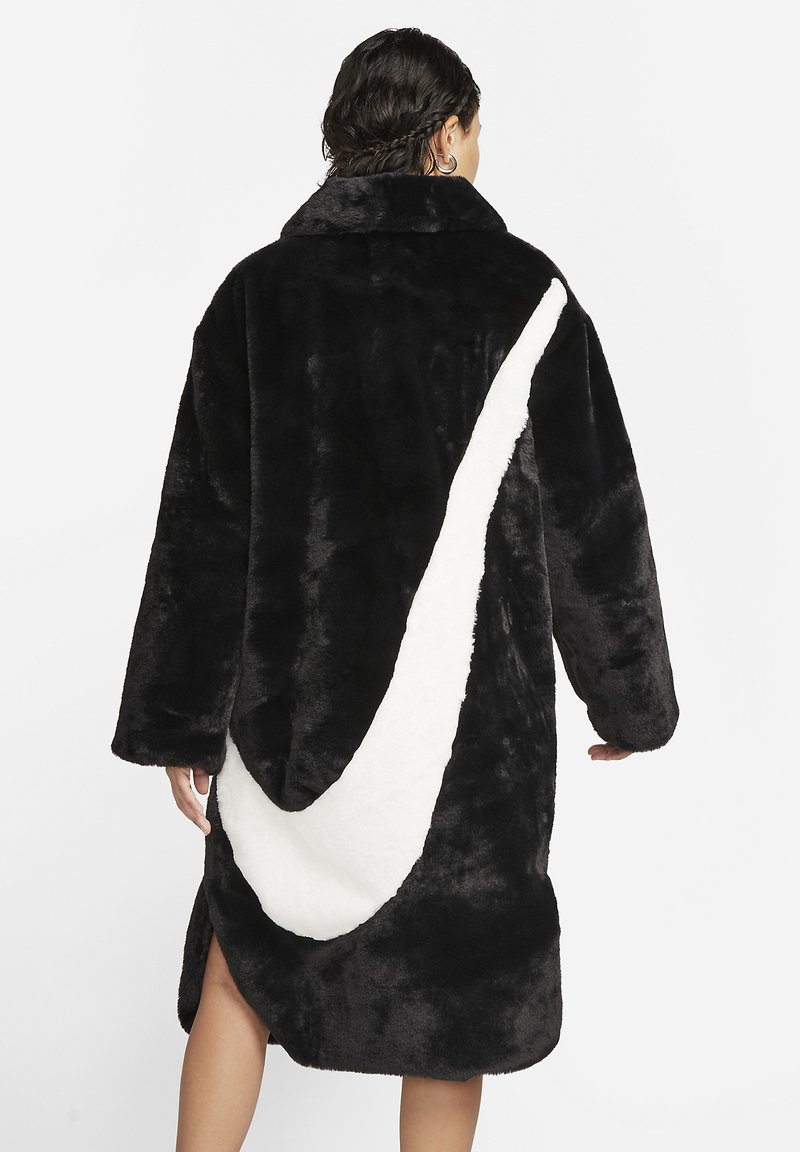 Abrigo de piel sintética negra con un alto cuello, que presenta un gran acento de swoosh blanco en la parte posterior y aberturas laterales para facilitar el movimiento.
