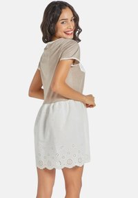 Joven mujer con cabello rizado sonríe mientras lleva una blusa beige y un vestido blanco de encaje, posando de espaldas. Fondo simple.