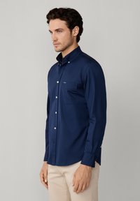 Chemise bleue marine à boutons, avec un col pointu, une poche frontale et des manches longues. Texture lisse, fabriquée en tissu léger. Présente un petit logo.