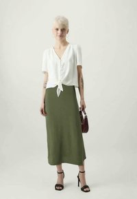 Blouse blanche à manches courtes avec un nœud à l'avant, associée à une longue jupe olive. Modèle tenant un petit sac à main bordeaux et portant des talons noirs.