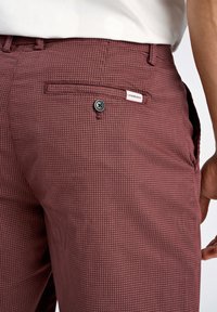 Pantaloni bordeaux texture con chiusura a bottone, tasche laterali e etichetta. Il tessuto presenta un motivo sottile.