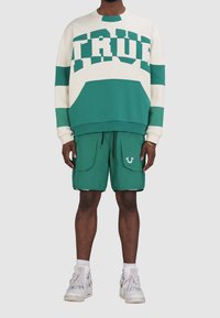 Zielono-biała bluza z napisem "TRUE", zestawiona z zielonymi szortami z dużymi kieszeniami i logo. Dopełnieniem są białe trampki.