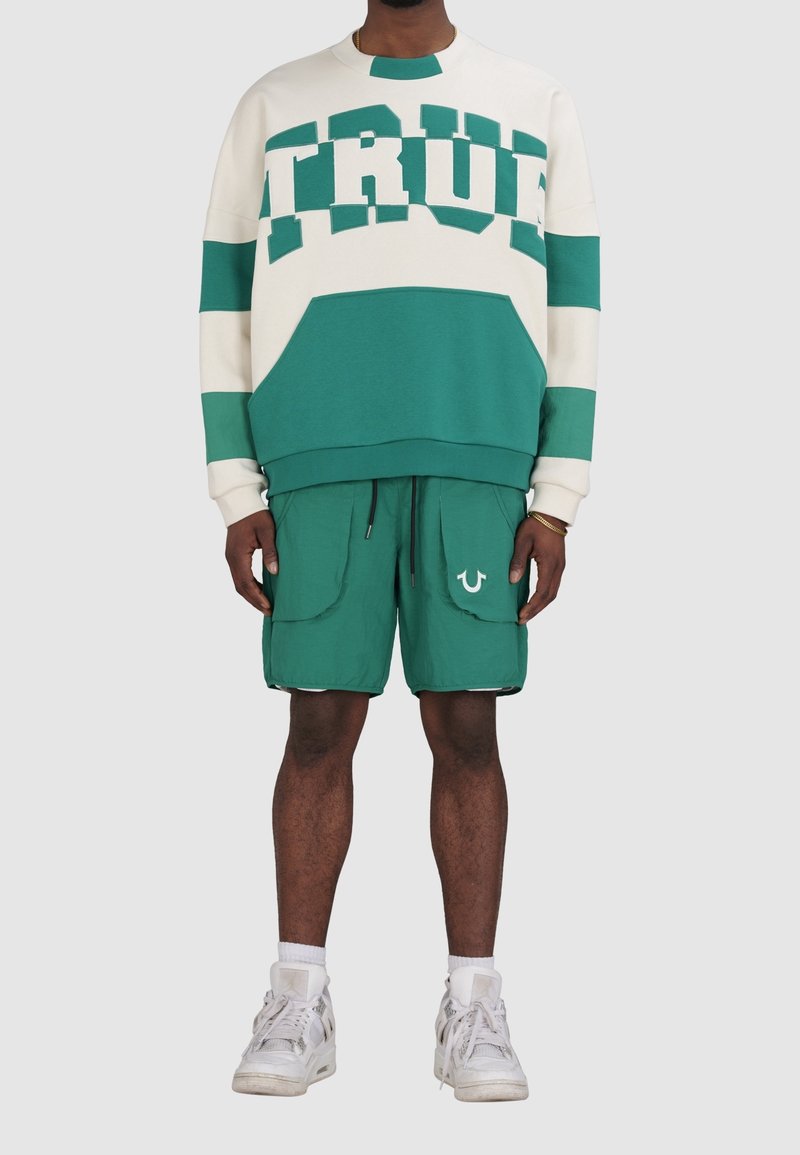 Zielono-biała bluza z napisem "TRUE", zestawiona z zielonymi szortami z dużymi kieszeniami i logo. Dopełnieniem są białe trampki.