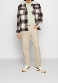 Chaqueta de franela a cuadros en blanco y negro con un collar blanco, combinada con una camiseta verde claro y pantalones beige. Zapatillas deportivas blancas y marrones.