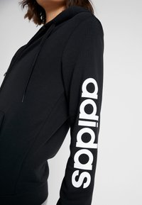 Persona vestida con una sudadera con cremallera negra y el logo blanco de "adidas" impreso verticalmente en la manga, frente a un fondo liso.