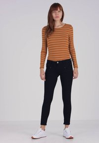 Top de manga larga a rayas marrón combinado con jeans ajustados negros. La modelo lleva zapatillas blancas con suelas naranjas, posando frente a un fondo claro.