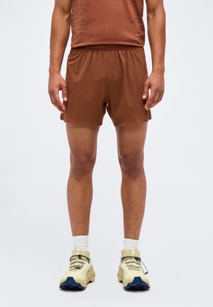 Mandlig model iført brune atletikshorts, en matchende brun top, hvide crew-sokker og beige løbesko med sorte snørebånd, der står på en hvid baggrund.