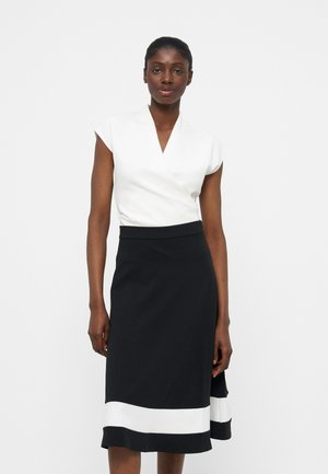 Vrouw draagt een mouwloze witte wikkeltop en een zwarte knie-lange rok met een brede witte streep nabij de zoom, staand tegen een effen achtergrond.