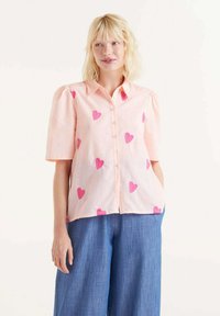 Camisa rosa de manga corta con hombros abullonados, botones en la parte delantera y patrones de corazones rosas dispersos. Combinada con pantalones de mezclilla azul holgados.