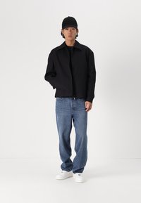 Črna zip-up jakna, jeans hlače in bele superge. Jakna ima brezkragelni dizajn; hlače so sproščenega kroja in svetlo modre barve.