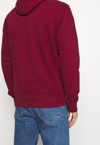 Sudadera burdeos hecha de tejido suave con un bolsillo canguro y puños de canalé. Presenta una capucha con cordón y un corte relajado.