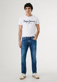 Bijela majica kratkih rukava s mornarsko plavim logotipom "Pepe Jeans London", uparen s plavim trapericama i bijelim tenisicama s bež akcentima.
