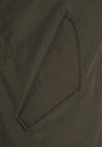 Donkergroen stof met een gladde textuur heeft een diagonale zak met contrasterende stiksels. De zakken hebben afgeronde randen en versterkte hoeken.