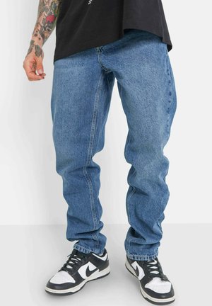 Vaqueros de mezclilla azul claro con un ajuste relajado, que presentan un ligero desgaste, costuras visibles y bolsillos laterales, combinados con zapatillas negras y blancas.
