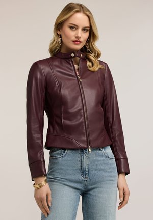 Kvinde iført en figursyet bordeaux læderjakke med lynlås, blå jeans, guldbøjler og hængende øreringe, stående foran en ensfarvet baggrund.