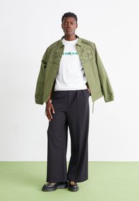 Πράσινο ελαφρύ μπουφάν με έντονες μαύρες ραφές, λευκό t-shirt με τεθαυγείς γραφικά, μαύρο παντελόνι φαρδύς γραμμής και μαύρες πλατφόρμες με ζωντανό σχέδιο ζώου.