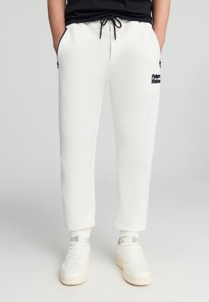 Persona che indossa pantaloni tuta bianchi con lacci neri, tasche laterali, testo nero sulla coscia e sneakers bianche su uno sfondo neutro.