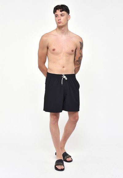 Pantaloni da bagno neri con coulisse bianca, vita elastica e tasche laterali. Modello che indossa sandali neri, in piedi su uno sfondo neutro.