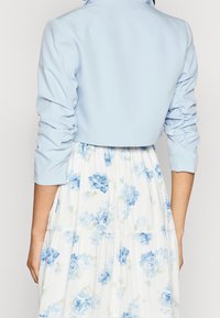 Veste courte bleu clair aux manches froncées portée par-dessus une robe blanche ornée d'un motif floral de roses bleues. La vue de dos révèle une texture de tissu lisse.